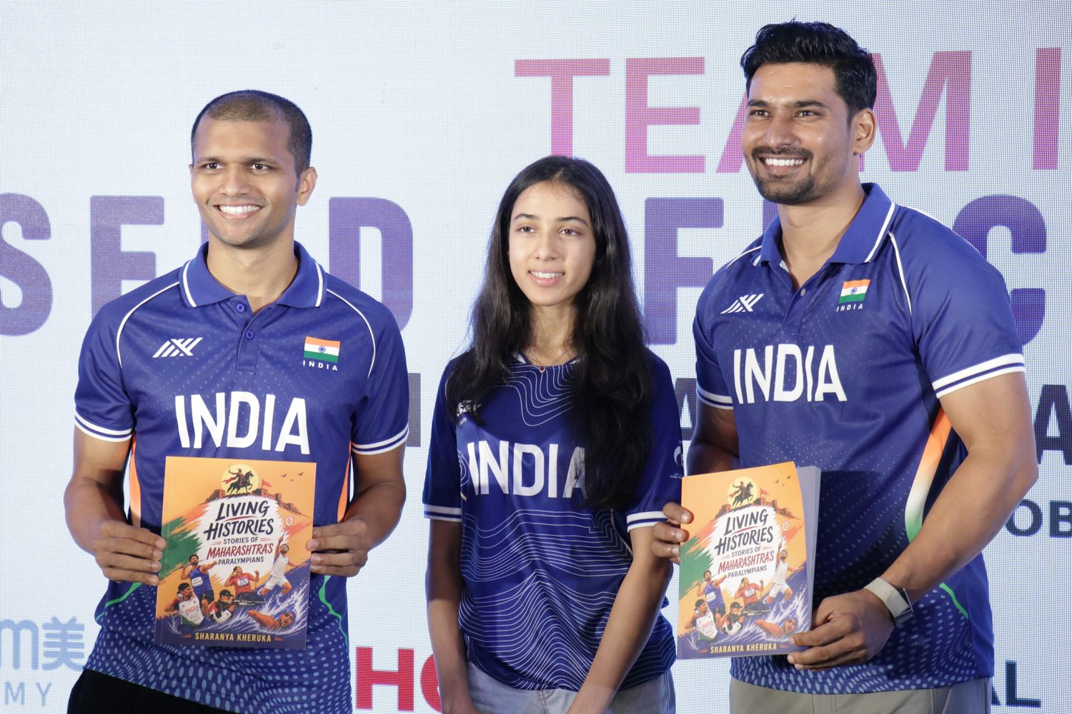 Sharanya Kheruka standing between two athletes in India jerseys, each holding the book